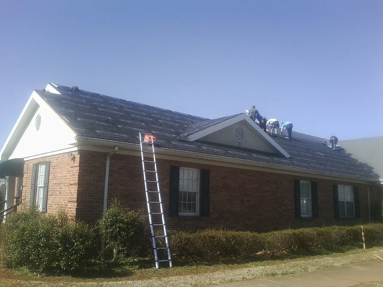 Crew tearing shingles off a brick ranch roof with a ladder set — Emergency Roof Repair work by our crew in Mystic